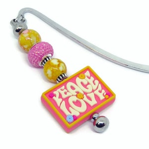 May include: A pink bookmark with a silver metal bookmark holder and a charm with the words "Peace Love" in white letters. The charm has yellow and pink accents and is attached to a string of beads.