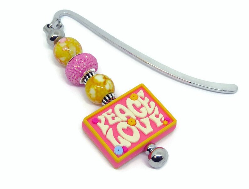May include: A pink bookmark with a silver metal bookmark holder and a charm with the words "Peace Love" in white letters. The charm has yellow and pink accents and is attached to a string of beads.