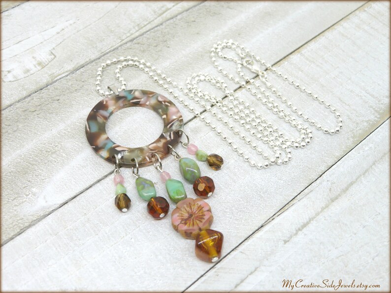 May include: A silver chain necklace with a large, round, multi-colored pendant. The pendant has a hole in the center and is decorated with a variety of beads, including pink, green, and brown.