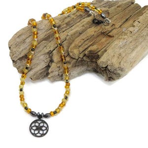 May include: A necklace featuring a circular pendant with a geometric flower-like pattern in black, suspended from a strand of translucent amber-colored beads interspersed with small, dark, metallic-toned beads and a silver-toned clasp.