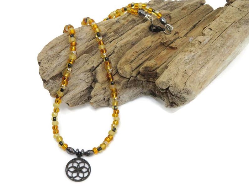 May include: A necklace featuring a circular pendant with a geometric flower-like pattern in black, suspended from a strand of translucent amber-colored beads interspersed with small, dark, metallic-toned beads and a silver-toned clasp.