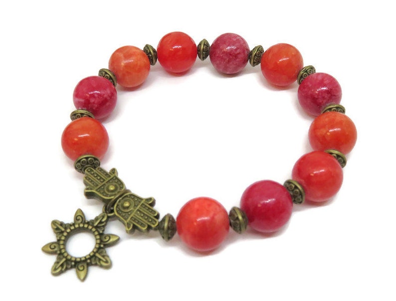 Pu&ograve; includere: Bracciale con perline rosse e arancioni e ciondoli color bronzo. Le perline rotonde sono separate da distanziatori color bronzo. I ciondoli includono un sole e una mano di Hamsa. Un gioiello colorato.