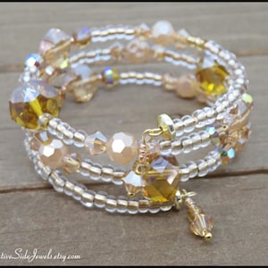 Puede incluir: Una pulsera envolvente de m&uacute;ltiples hilos con cuentas transparentes y doradas, con cristales facetados de color &aacute;mbar y melocot&oacute;n. La pulsera tiene un cierre dorado y un dije colgante. La joya se exhibe sobre una superficie de madera.