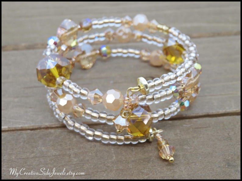 Puede incluir: Una pulsera envolvente de m&uacute;ltiples hilos con cuentas transparentes y doradas, con cristales facetados de color &aacute;mbar y melocot&oacute;n. La pulsera tiene un cierre dorado y un dije colgante. La joya se exhibe sobre una superficie de madera.