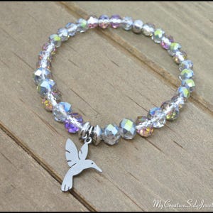 May include: A stretch bracelet featuring faceted, iridescent clear beads with subtle purple and blue highlights. A silver-toned metal hummingbird charm hangs from the center of the circular bead strand. The text MyCreativeSideJewels.etsy.com is visible.