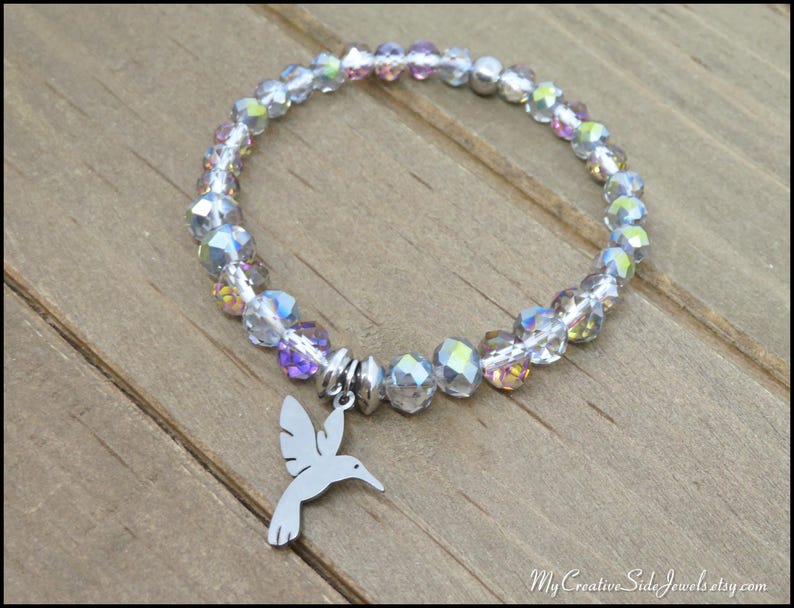 May include: A stretch bracelet featuring faceted, iridescent clear beads with subtle purple and blue highlights. A silver-toned metal hummingbird charm hangs from the center of the circular bead strand. The text MyCreativeSideJewels.etsy.com is visible.