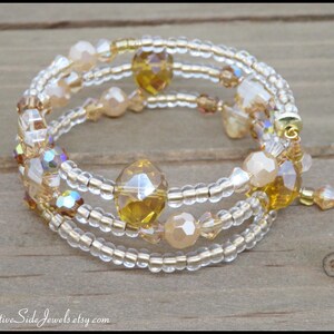 Puede incluir: Una pulsera envolvente de m&uacute;ltiples hilos con cuentas transparentes y doradas, con detalles de cristal iridiscente y &aacute;mbar. La pulsera est&aacute; enrollada sobre s&iacute; misma, mostrando su dise&ntilde;o en capas. La pulsera est&aacute; sobre una superficie de madera.
