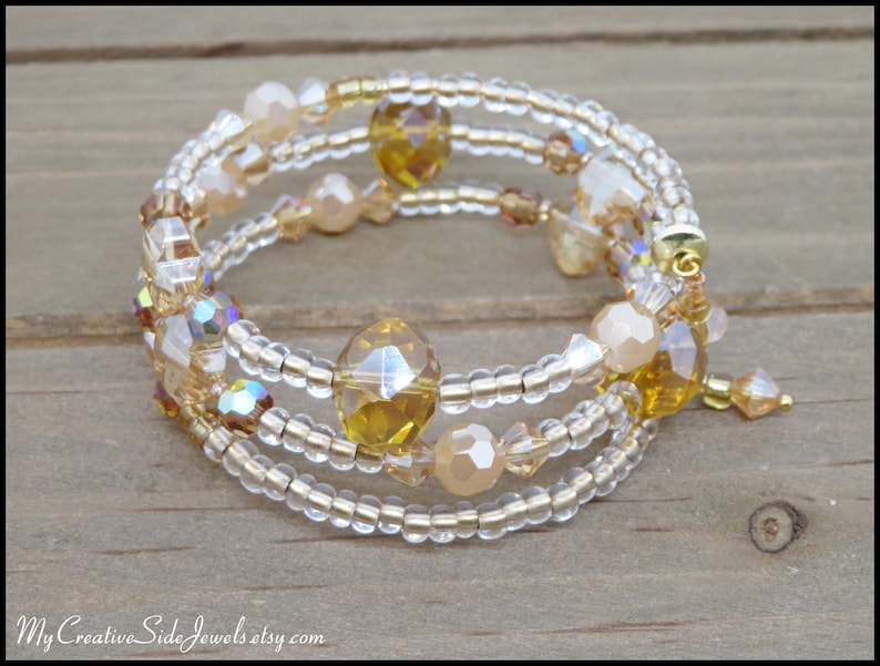 Puede incluir: Una pulsera envolvente de m&uacute;ltiples hilos con cuentas transparentes y doradas, con detalles de cristal iridiscente y &aacute;mbar. La pulsera est&aacute; enrollada sobre s&iacute; misma, mostrando su dise&ntilde;o en capas. La pulsera est&aacute; sobre una superficie de madera.