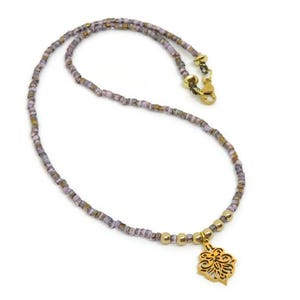 May include: A delicate necklace featuring small, square, lavender and gold beads. The necklace has a gold-colored clasp and a decorative gold pendant with an openwork floral design. The necklace is displayed on a white background.