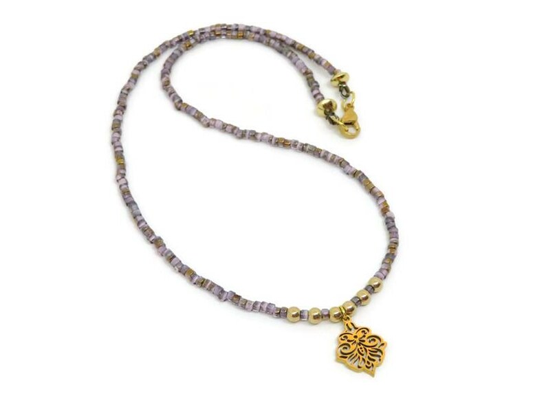 May include: A delicate necklace featuring small, square, lavender and gold beads. The necklace has a gold-colored clasp and a decorative gold pendant with an openwork floral design. The necklace is displayed on a white background.