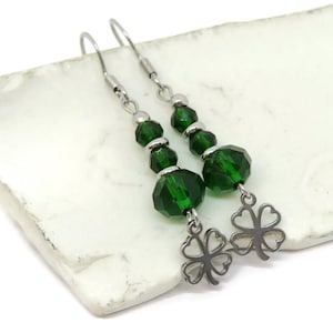 May include: A pair of dangle earrings featuring faceted emerald green beads and silver-tone clover charms. The earrings have a hook closure and are displayed on a white, textured surface. The jewelry is suitable for St. Patrick's Day.