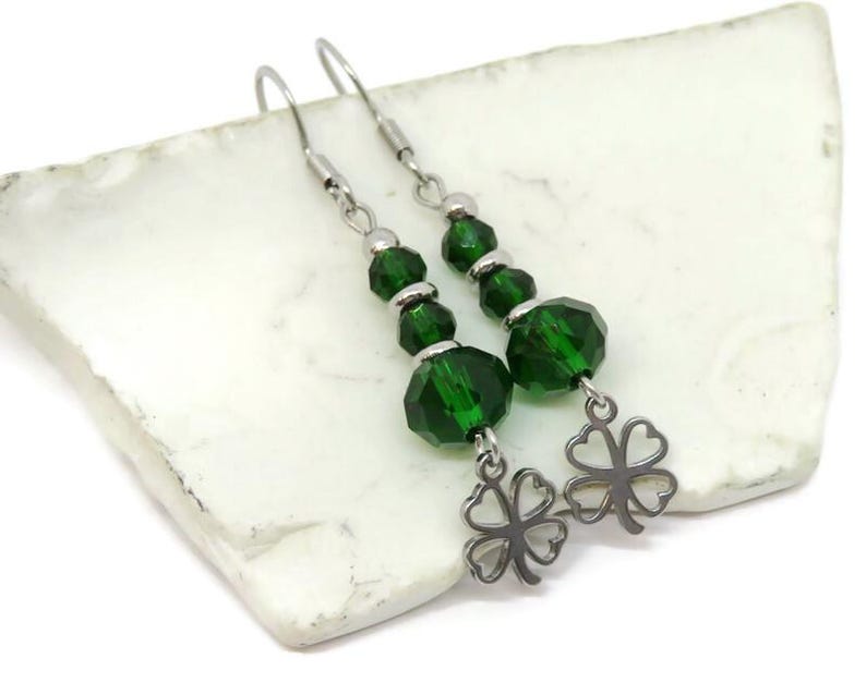 May include: A pair of dangle earrings featuring faceted emerald green beads and silver-tone clover charms. The earrings have a hook closure and are displayed on a white, textured surface. The jewelry is suitable for St. Patrick's Day.