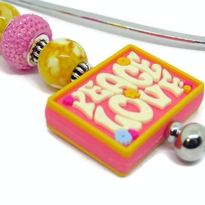 May include: A pink and yellow bookmark with a silver metal bookmark holder. The bookmark has a square charm with the words "Peace Love" in white letters. The charm is decorated with small pink, yellow, and blue flowers.