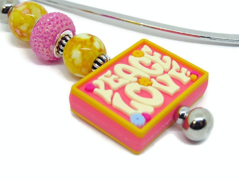 May include: A pink and yellow bookmark with a silver metal bookmark holder. The bookmark has a square charm with the words "Peace Love" in white letters. The charm is decorated with small pink, yellow, and blue flowers.