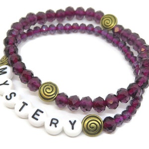 May include: A double-strand bracelet with purple glass beads and gold accents. The bracelet has the word "MYSTERY" spelled out in white beads.