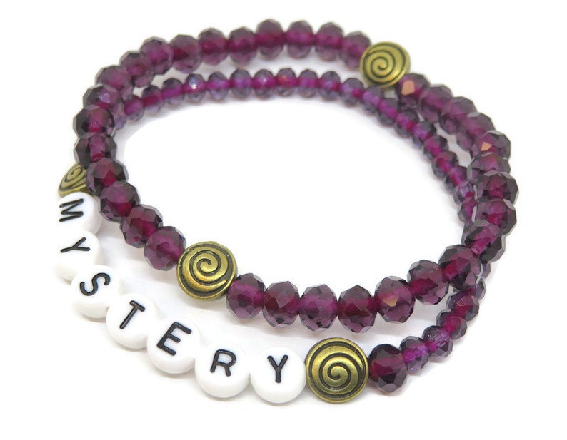 May include: A double-strand bracelet with purple glass beads and gold accents. The bracelet has the word "MYSTERY" spelled out in white beads.