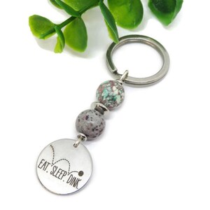 May include: A silver keychain with two speckled beads and a round charm. The charm is engraved with the words "EAT. SLEEP. DINK" and a pickleball graphic. The keychain has a silver ring for attaching keys.