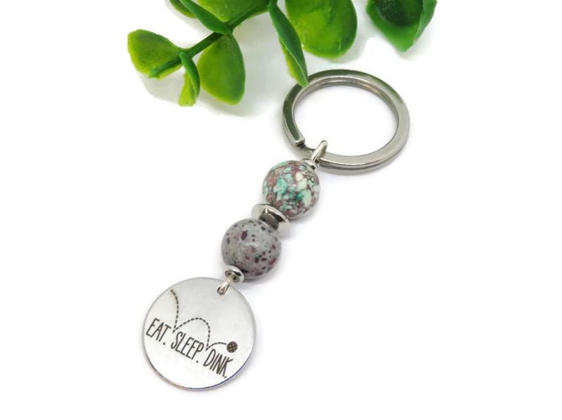 May include: A silver keychain with two speckled beads and a round charm. The charm is engraved with the words "EAT. SLEEP. DINK" and a pickleball graphic. The keychain has a silver ring for attaching keys.