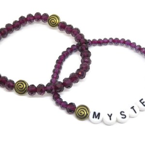 May include: Two purple crystal beaded bracelets with gold spiral charms. One bracelet has the word "MYSTERY" spelled out in white beads.