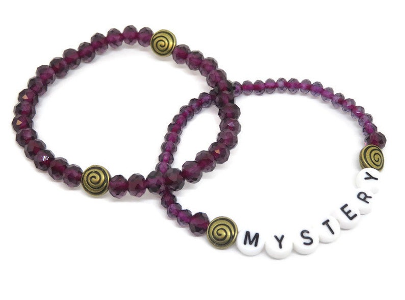 May include: Two purple crystal beaded bracelets with gold spiral charms. One bracelet has the word "MYSTERY" spelled out in white beads.