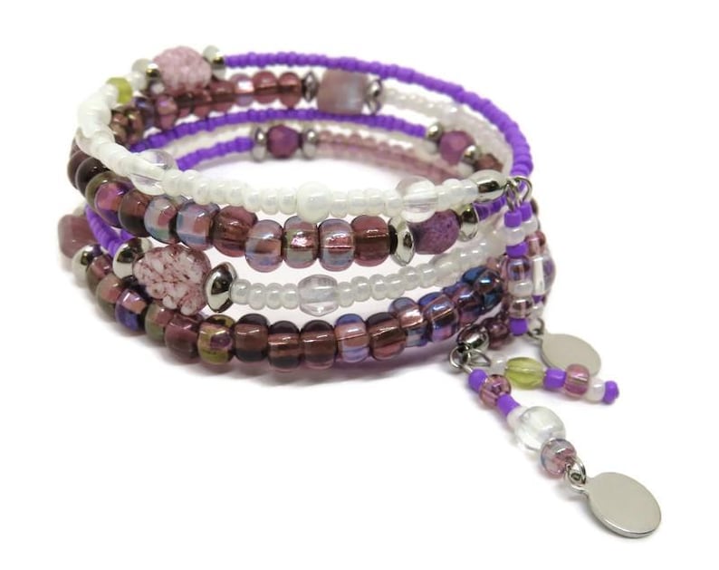 May include: A multi-strand wrap bracelet with purple, white, and burgundy beads. The bracelet features a mix of round and square beads, with dangling charms. The beads are strung on a wire, creating a spiral design.