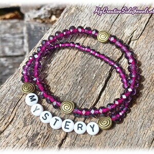May include: A purple crystal beaded bracelet with a gold charm and white letter beads spelling out the word "MYSTERY".