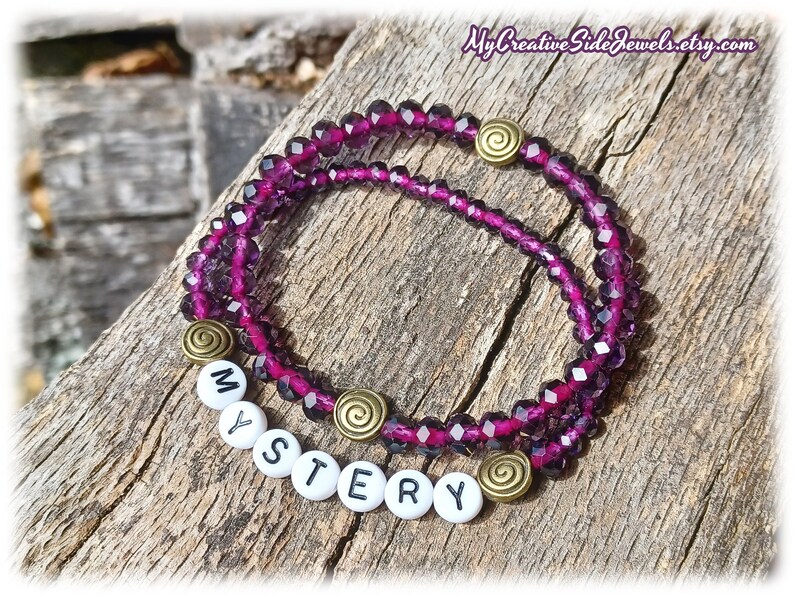 May include: A purple crystal beaded bracelet with a gold charm and white letter beads spelling out the word "MYSTERY".