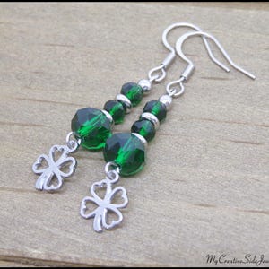 May include: A pair of dangle earrings featuring faceted emerald green beads and silver-tone four-leaf clover charms. The earrings have silver-tone ear wires and small silver beads. The jewelry is displayed on a wooden surface.