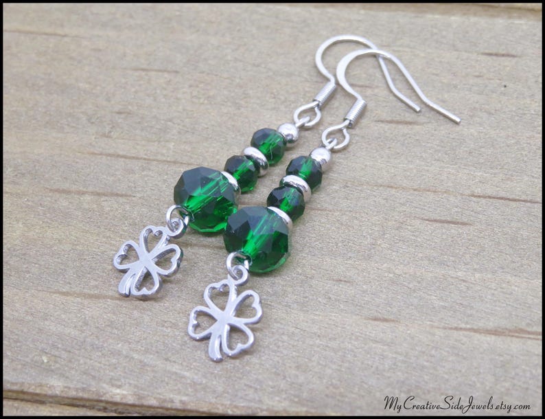 May include: A pair of dangle earrings featuring faceted emerald green beads and silver-tone four-leaf clover charms. The earrings have silver-tone ear wires and small silver beads. The jewelry is displayed on a wooden surface.