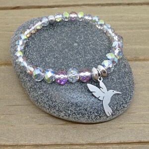 May include: A stretch bracelet featuring faceted, iridescent clear and light purple crystal beads with a silver-toned metal hummingbird charm. The charm is attached to the bracelet by a smooth, round silver-toned metal bead spacer.