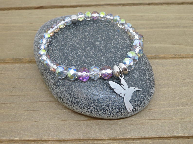May include: A stretch bracelet featuring faceted, iridescent clear and light purple crystal beads with a silver-toned metal hummingbird charm. The charm is attached to the bracelet by a smooth, round silver-toned metal bead spacer.