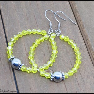 May include: A pair of yellow crystal hoop earrings with silver metal accents. The earrings are made with small, faceted crystals and have a simple, elegant design.