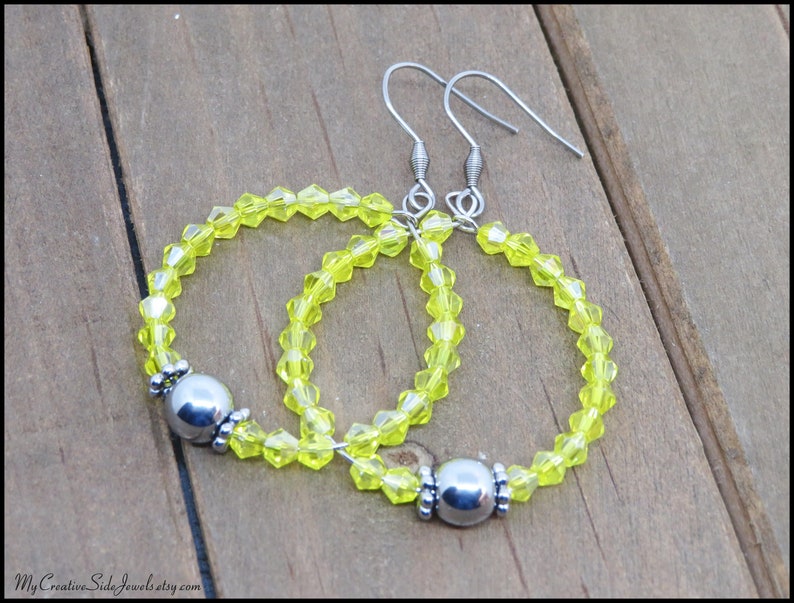 May include: A pair of yellow crystal hoop earrings with silver metal accents. The earrings are made with small, faceted crystals and have a simple, elegant design.