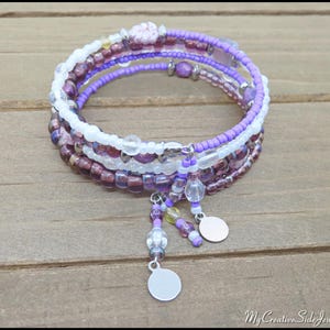 May include: A multi-strand wrap bracelet featuring a mix of purple, white, and clear beads. The bracelet has dangling charms and is displayed on a wooden surface. The bracelet is a jewelry item.