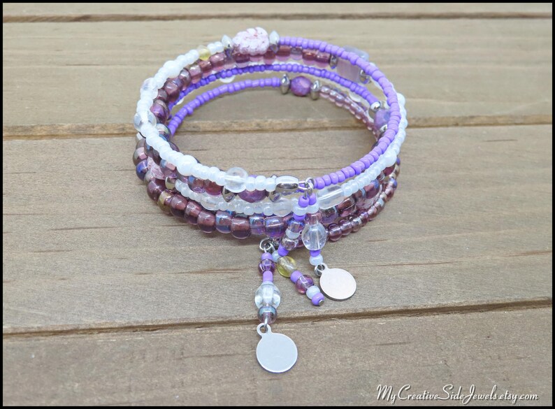 May include: A multi-strand wrap bracelet featuring a mix of purple, white, and clear beads. The bracelet has dangling charms and is displayed on a wooden surface. The bracelet is a jewelry item.