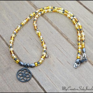 May include: A beaded necklace featuring a circular, openwork metal pendant with a geometric flower pattern. The strand consists of alternating translucent amber, yellow, and metallic gray seed beads, finished with a dark metal lobster claw clasp.