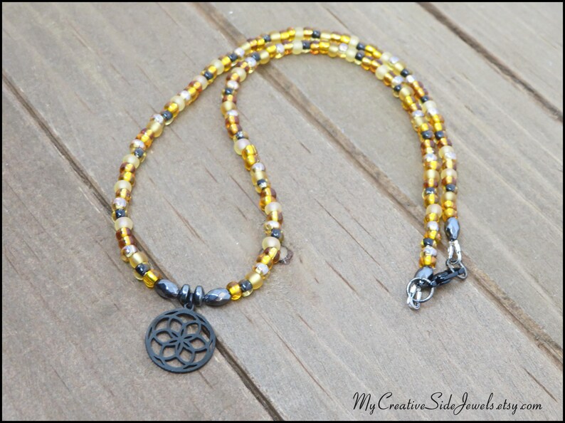 May include: A beaded necklace featuring a circular, openwork metal pendant with a geometric flower pattern. The strand consists of alternating translucent amber, yellow, and metallic gray seed beads, finished with a dark metal lobster claw clasp.