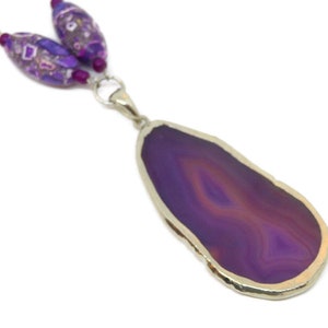 May include: A purple agate pendant necklace with a silver setting. The pendant is a large, freeform slice of agate with a natural edge. The necklace is made with purple and white glass beads.