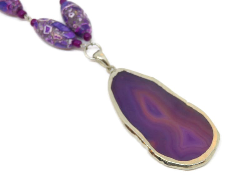 May include: A purple agate pendant necklace with a silver setting. The pendant is a large, freeform slice of agate with a natural edge. The necklace is made with purple and white glass beads.