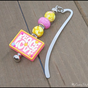 May include: A pink and yellow bookmark with the words "Peace Love" on a pink rectangle charm. The bookmark has a silver metal bookmark with a chain of pink and yellow beads.