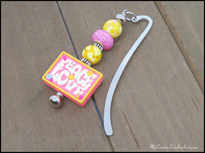 May include: A pink and yellow bookmark with the words "Peace Love" on a pink rectangle charm. The bookmark has a silver metal bookmark with a chain of pink and yellow beads.