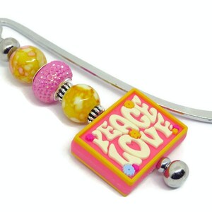 May include: A silver bookmark with a pink and yellow charm that says "Peace Love" in white letters. The charm has small yellow flowers around the border. The bookmark has three beads: one yellow with white speckles, one pink with glitter, and one yellow with white speckles.