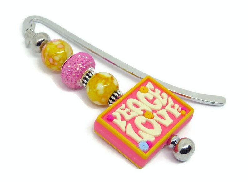 May include: A silver bookmark with a pink and yellow charm that says "Peace Love" in white letters. The charm has small yellow flowers around the border. The bookmark has three beads: one yellow with white speckles, one pink with glitter, and one yellow with white speckles.