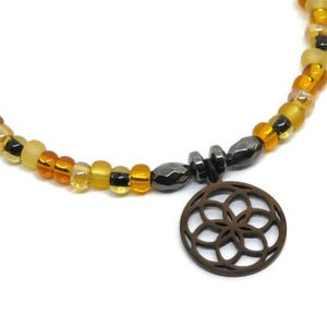 May include: A beaded necklace featuring a circular pendant with a geometric flower-like cutout design. The necklace is composed of alternating translucent and opaque beads in shades of amber, yellow, and black, accented by dark metallic beads.