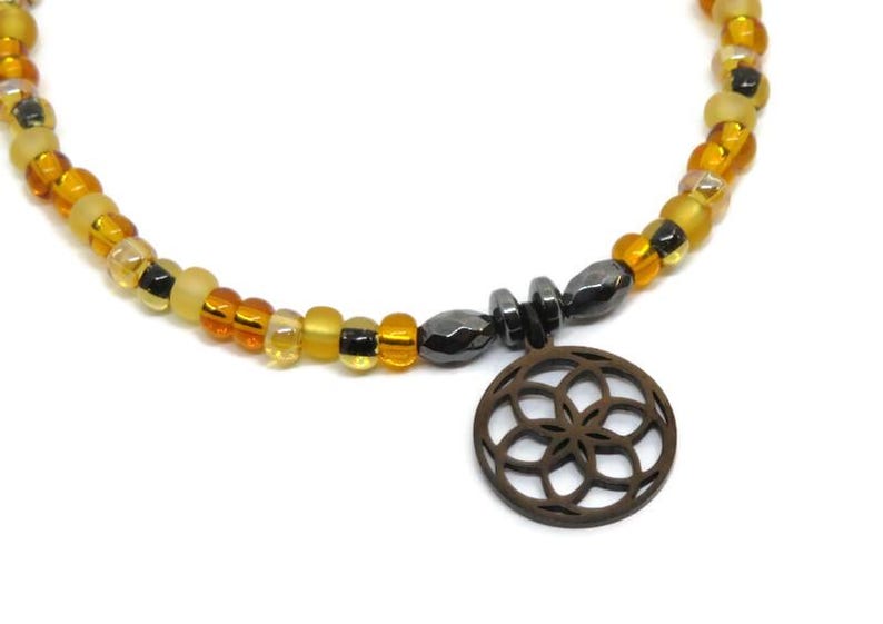 May include: A beaded necklace featuring a circular pendant with a geometric flower-like cutout design. The necklace is composed of alternating translucent and opaque beads in shades of amber, yellow, and black, accented by dark metallic beads.