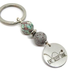 May include: Silver keychain with a round charm that says "EAT. SLEEP. DINK." The keychain has two gray and turquoise beads. The charm has a graphic of a bouncing ball.
