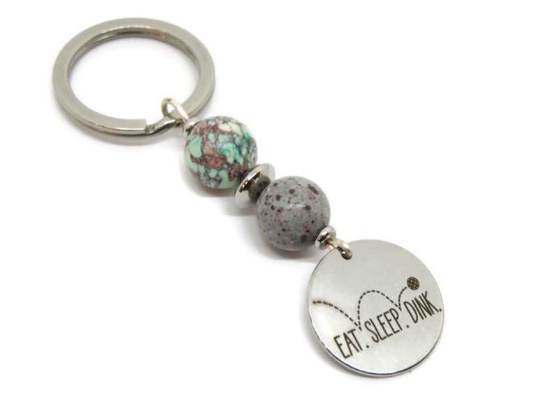 May include: Silver keychain with a round charm that says "EAT. SLEEP. DINK." The keychain has two gray and turquoise beads. The charm has a graphic of a bouncing ball.