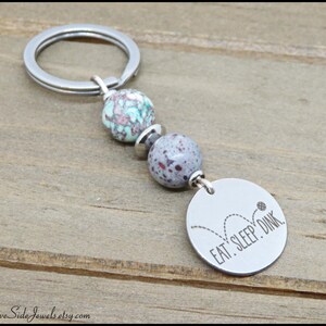 May include: A silver keychain with a round tag that says "EAT. SLEEP. DINK." The keychain has two speckled beads in shades of blue and purple. The keychain is on a wooden surface.