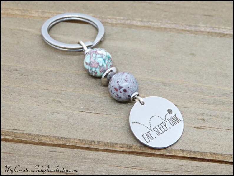 May include: A silver keychain with a round tag that says "EAT. SLEEP. DINK." The keychain has two speckled beads in shades of blue and purple. The keychain is on a wooden surface.