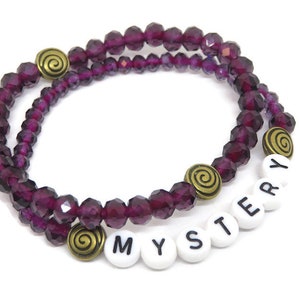 May include: Purple crystal bead bracelet with gold accents and white letter beads spelling "MYSTERY".